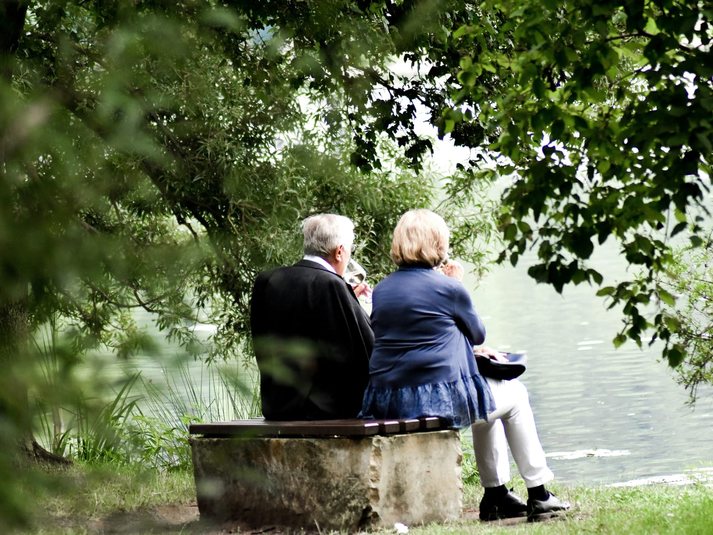 Casal de idosos sentado em um banco à beira de um lago, sob as árvores